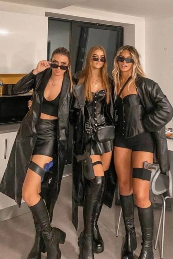 This image is of a girl group dressed up as detectives for halloween. They are wearing all black and long trenchcoats.