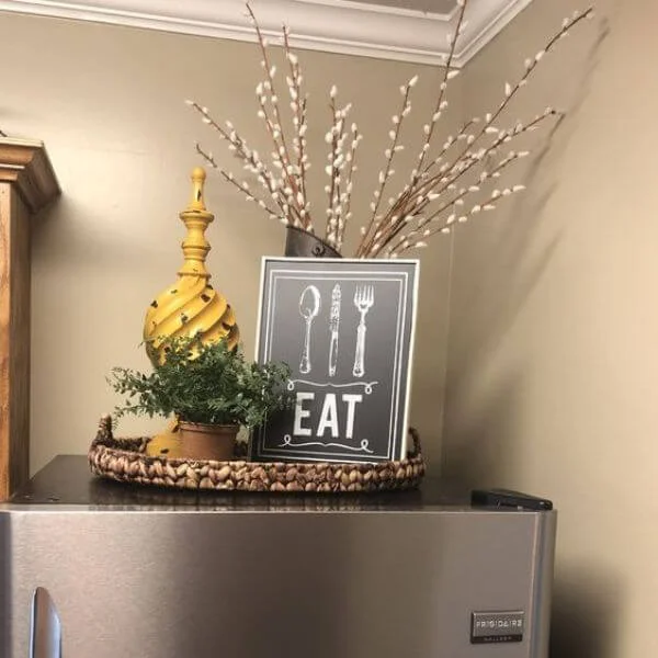 This image is of a home decor look on top of the fridge trhat has a sign that says eat on it. There are fake plants to compliment it.